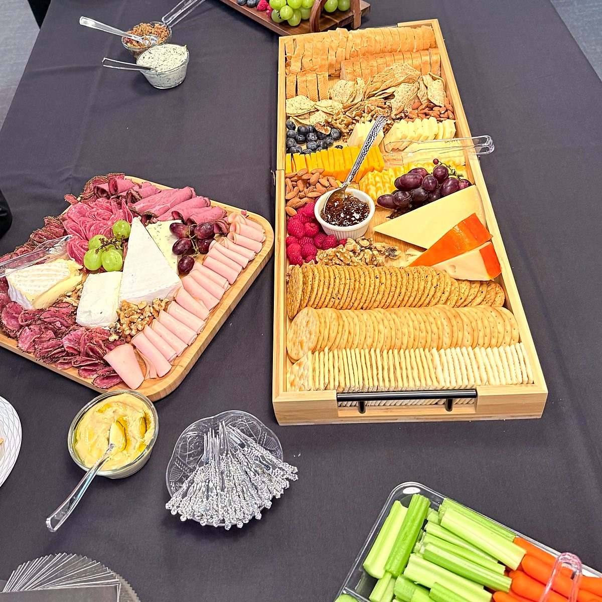 Office event spread with a charcuterie board, a cheese and cracker tray, hummus and a vegetable platter on a dark tablecloth
