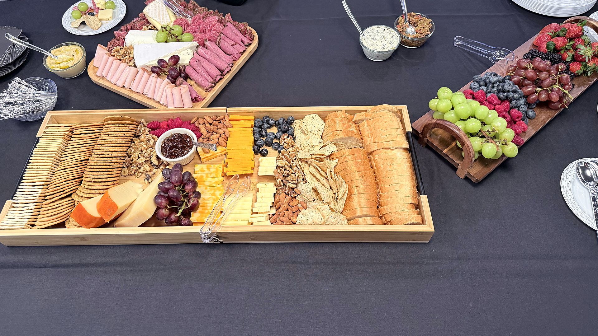 Thyme Catering grazing table on a black linen with a long wooden tray of crackers, cheeses, nuts, grapes and sliced baguette, a charcuterie board and a fruit board of berries and grapes