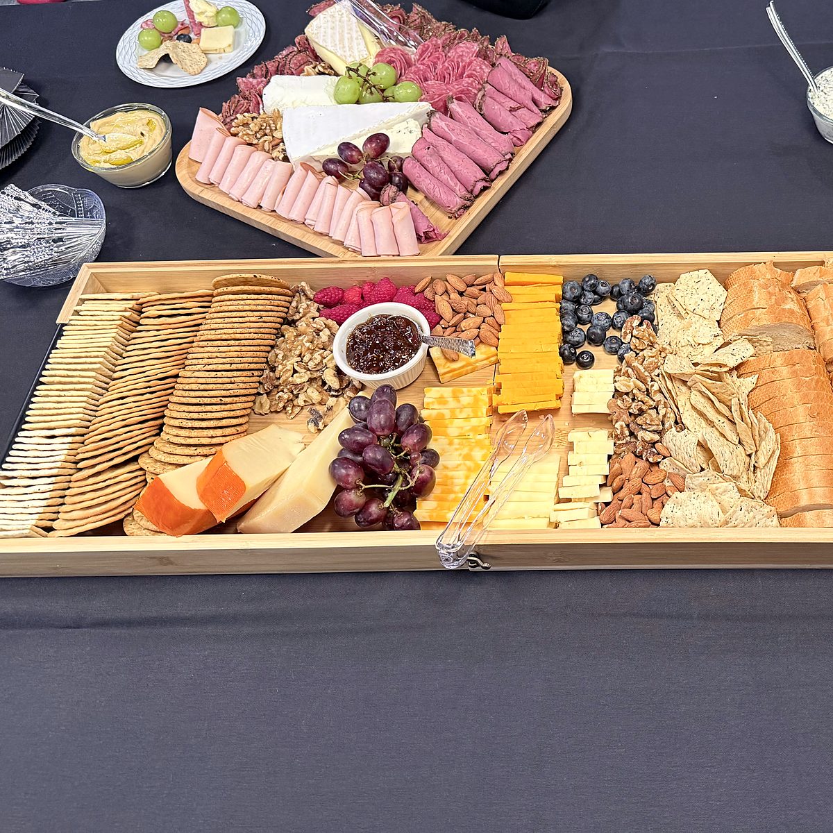 Close-up of a Thyme Catering charcuterie board and a wooden tray of crackers, cheddar, gouda, grapes, nuts and fig jam on a black tablecloth